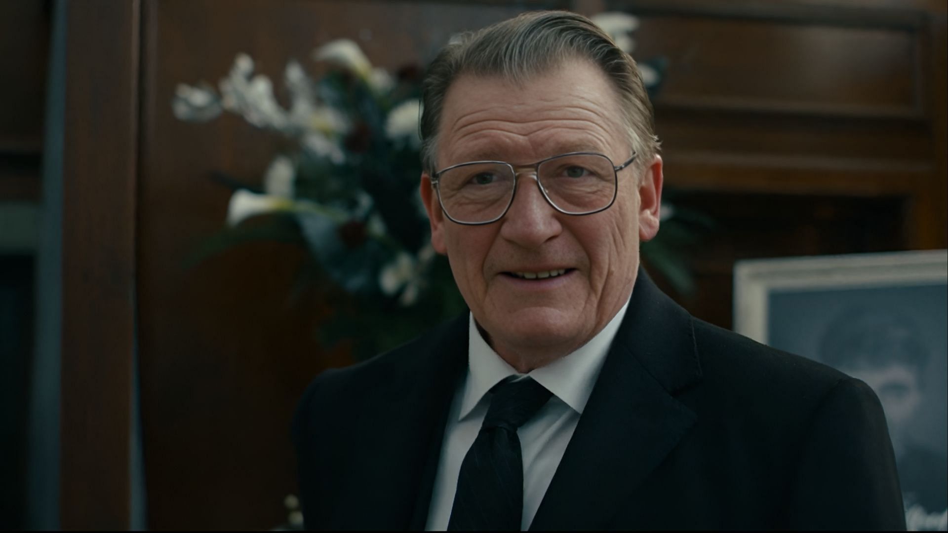 A rare moment of charm: Richie flashes a smile, but the room behind him speaks of funerals and facades. (Image via Paramount)